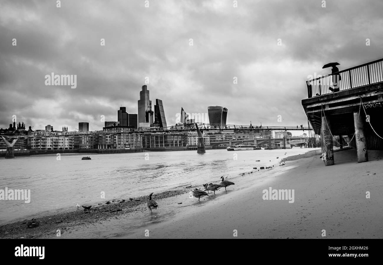 Photographie urbaine en noir et blanc à Londres : la Tamise à marée basse avec un ciel orageux au-dessus de la ville de Londres. Banque D'Images