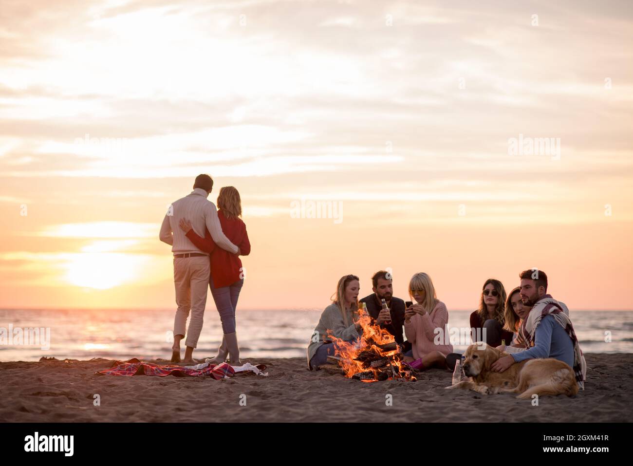 Jeune couple avec des amis autour de feu de camp sur la plage au coucher du soleil la consommation de bière Banque D'Images