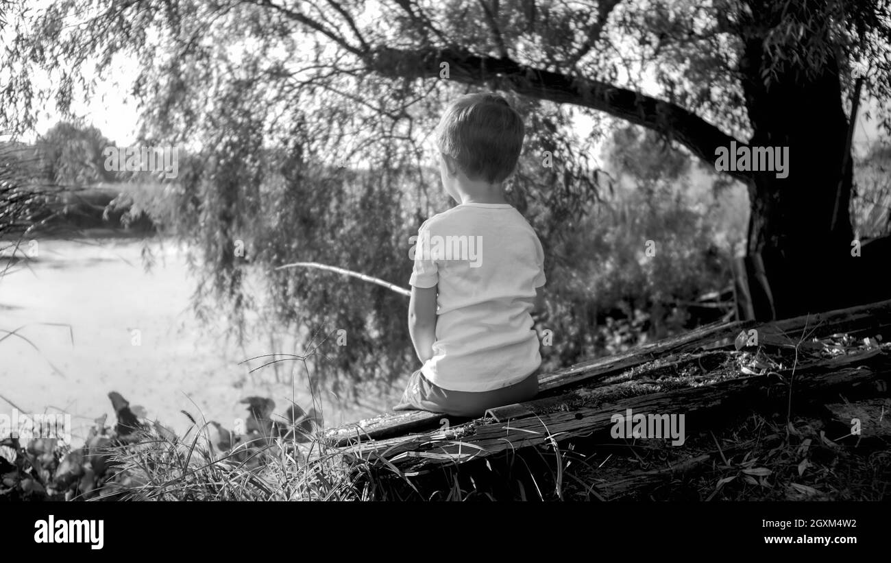 Photo noir et blanc de petit garçon avec canne à pêche en bois accrochant du poisson sur le lac. Banque D'Images