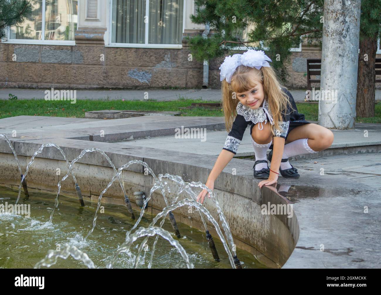 Une belle petite fille dans une robe d'école joue avec les jets de la fontaine. Banque D'Images