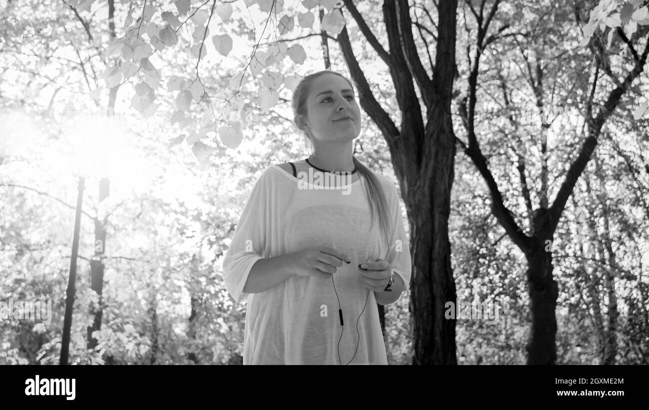 Photo en noir et blanc d'une jeune femme souriante en tenue de sport avec écouteurs au parc ensoleillé. Banque D'Images