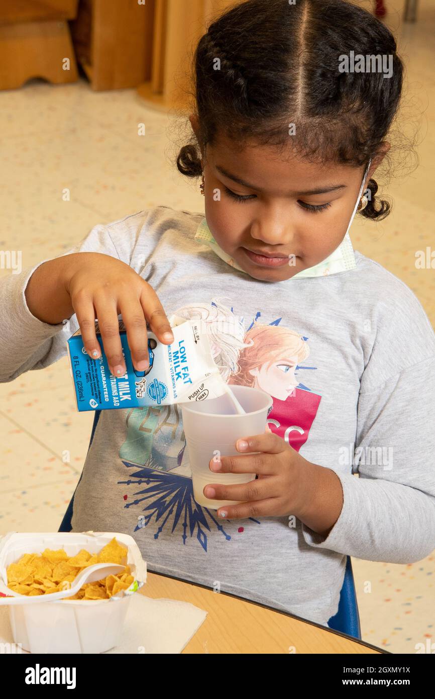 Éducation préscolaire 3-4 ans jeune fille versant du lait dans ses céréales au petit déjeuner, masque de visage vs Covid-19 tiré vers le bas pour le temps de repas Banque D'Images