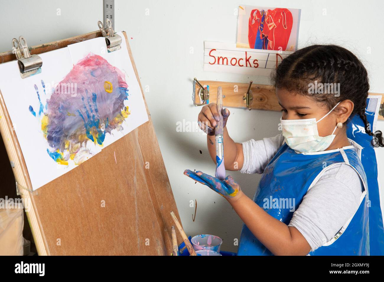 Éducation préscolaire 4-5 ans jeune fille dans la peinture de smock au easel, brossant la peinture sur sa propre main, portant un masque facial pour protéger contre Covid-19 Banque D'Images