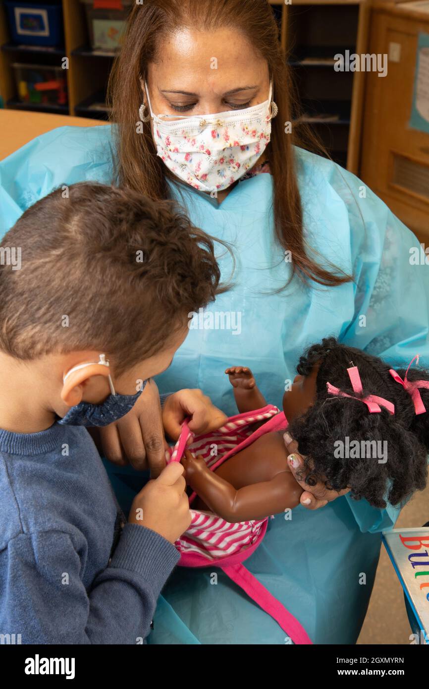 Éducation préscolaire 3-4 ans une enseignante tient la poupée comme le garçon travaille sur la tenue, la tâche de moteur fine. Les deux portent des masques de protection contre Covid-19 Banque D'Images