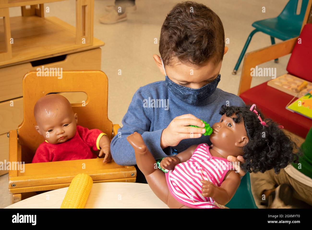 Éducation les jeunes de 3-4 ans préscolaires prétendent jouer dans la zone familiale un garçon nourrissant une poupée avec de la nourriture de jeu, portant un masque facial en raison de Covid-19 Banque D'Images
