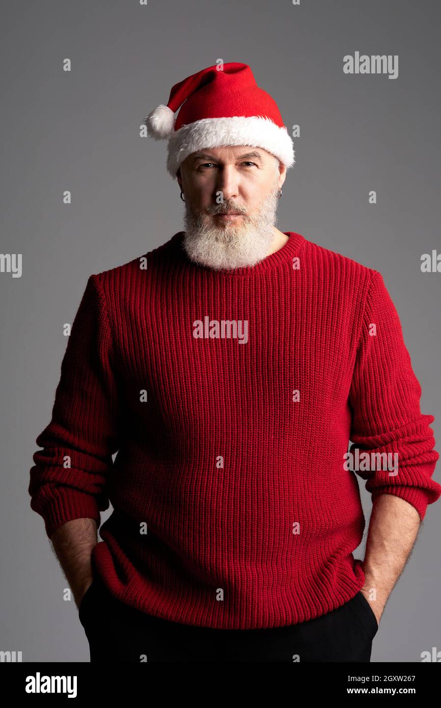Père Noël élégant. Homme d'âge moyen avec barbe portant un chapeau de Noël et un chandail rouge regardant l'appareil photo tout en se tenant isolé sur fond gris Banque D'Images