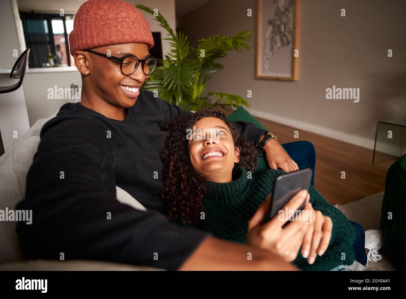 Couple multiethnique élégant souriant en regardant le téléphone portable.Appartement moderne sur le canapé Banque D'Images