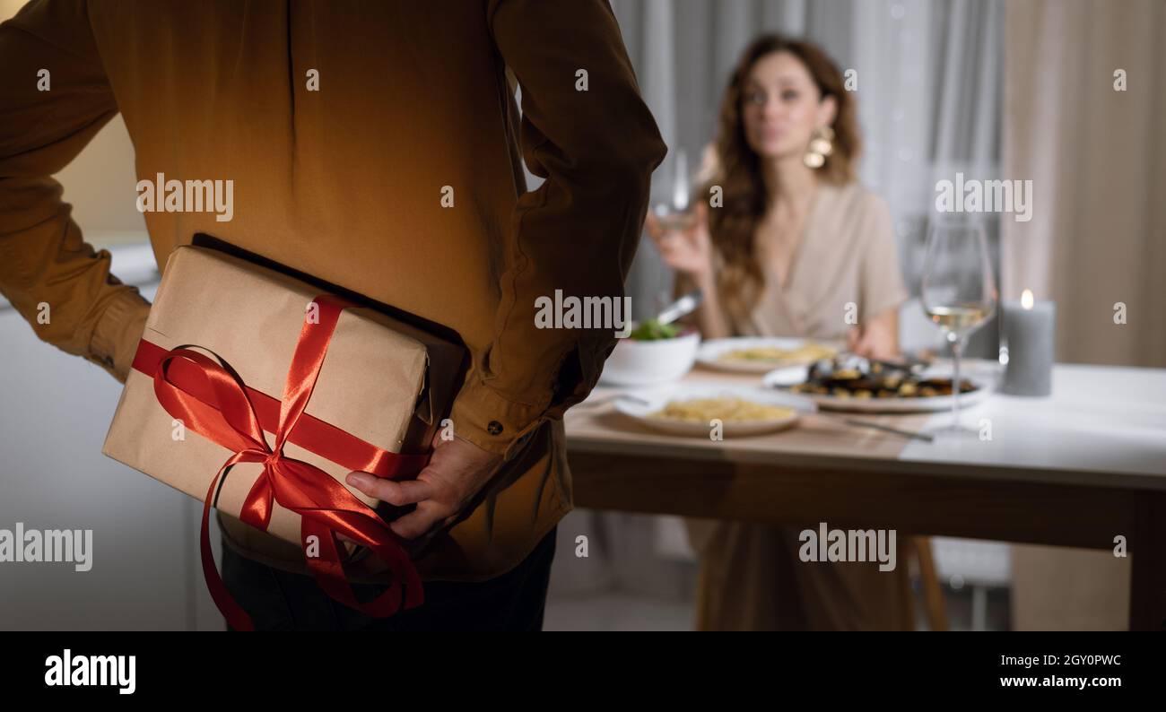la femme est assise à la table dans une robe, l'homme cache la boîte derrière son dos.Concept cadeau pour la Saint-Valentin, l'anniversaire, Banque D'Images