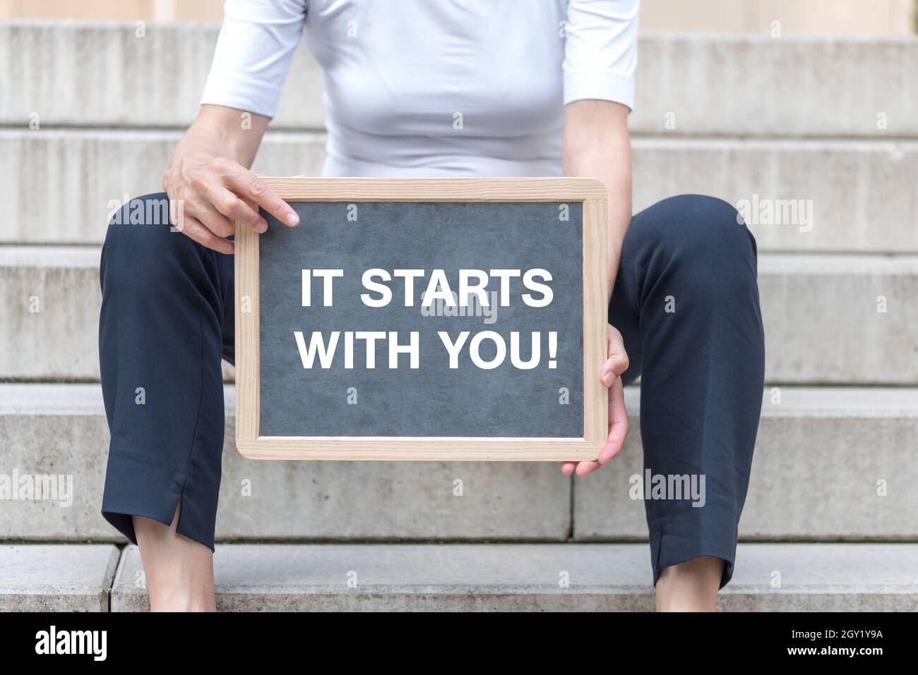 Femme d'âge moyen assise dans les escaliers et tenant un tableau noir avec le texte « ça commence avec vous ! » Banque D'Images