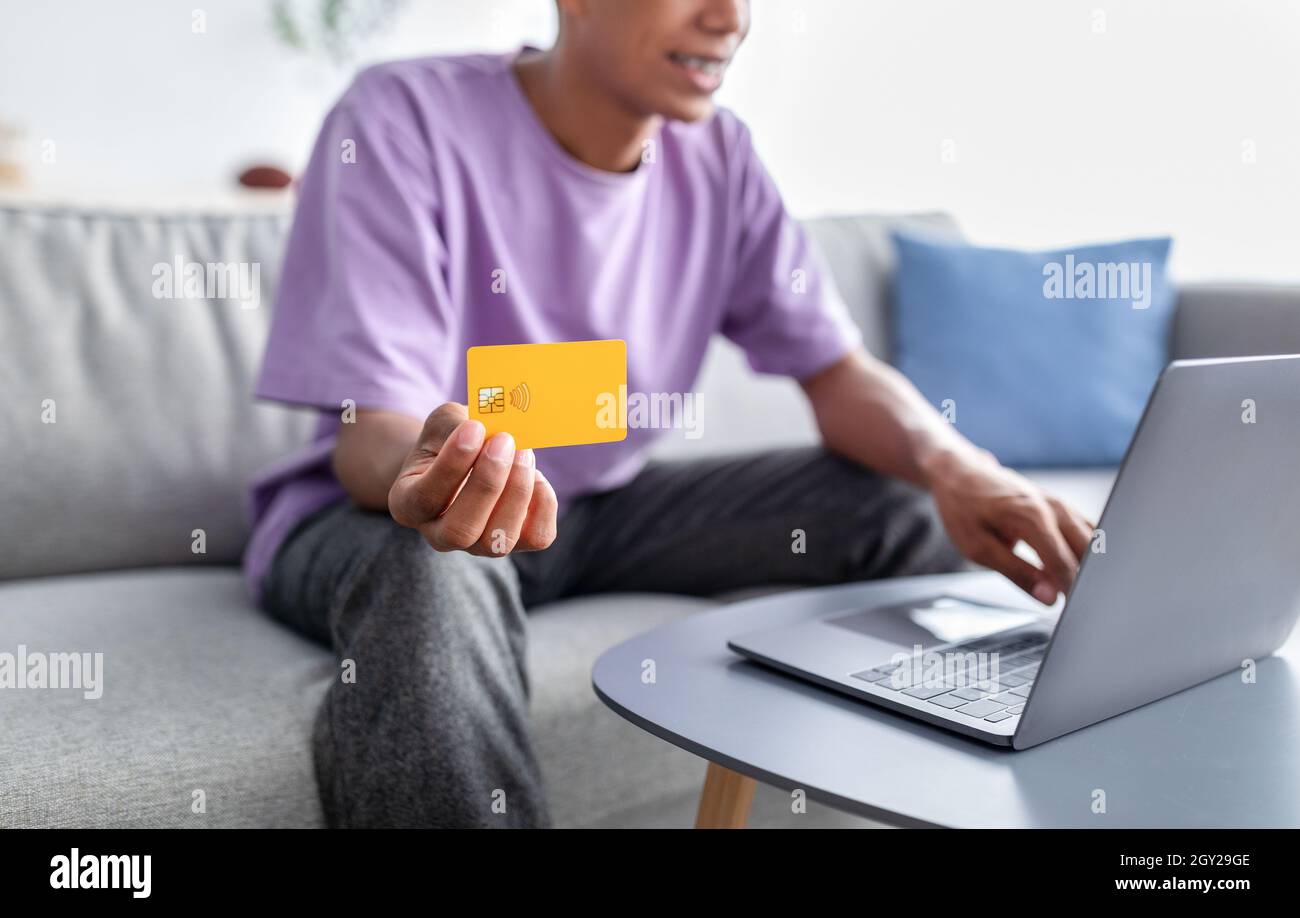 Concept d'achat à distance.Adolescent noir méconnaissable avec ordinateur portable montrant une carte de crédit, commandant des produits en ligne à la maison Banque D'Images