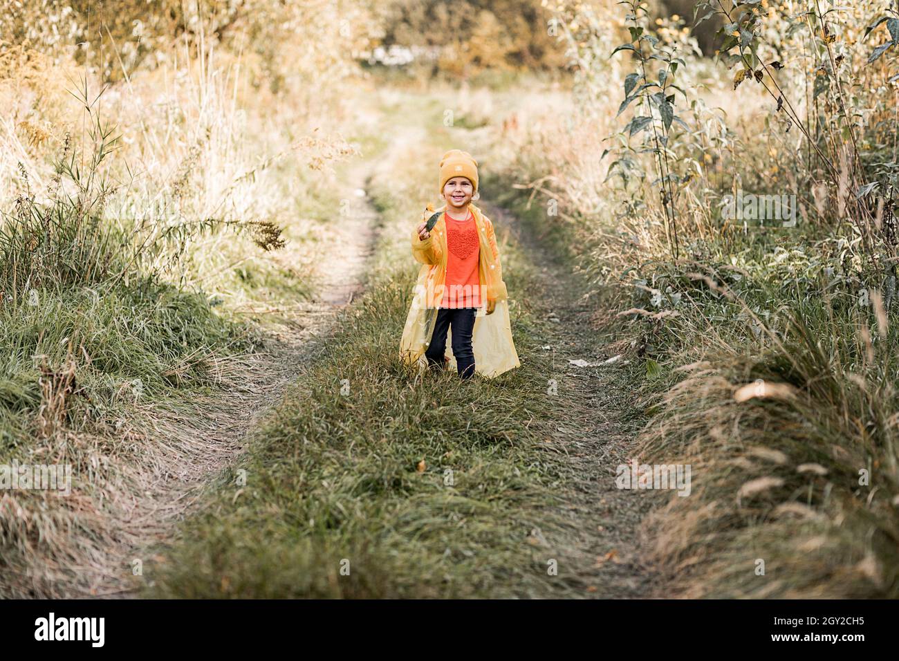 photo d'une petite fille drôle en chapeau jaune et imperméable marchant le long de la route parmi l'herbe haute et souriant Banque D'Images