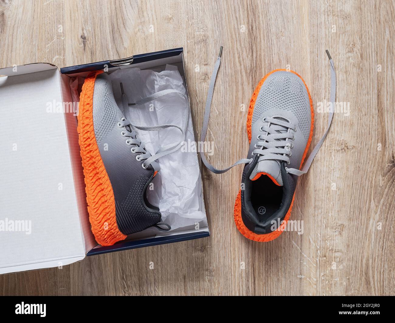 Une sneaker dans la boîte en carton ouverte et l'autre près du sol.Nouvelle paire de sneakers en tissu gris avec semelle rainurée orange.Chaussures en mesh élégantes. Banque D'Images
