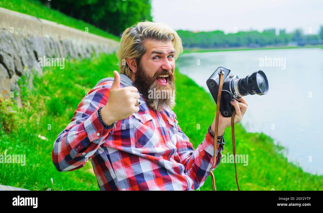 Homme barbu prenant des photos avec un appareil photo sur la rivière.Passe-temps et voyages.Homme souriant avec un appareil photo numérique professionnel. Banque D'Images