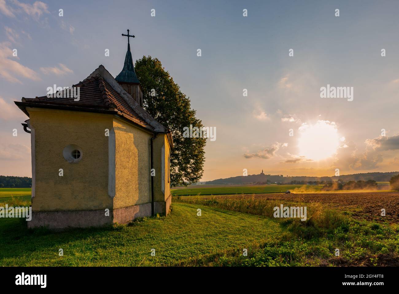 Scène typique de travail agricole rurale bavaroise avec petite chapelle, terrain de labour de tracteur et église de pèlerinage au sommet de la colline en arrière-plan en soirée sunlig Banque D'Images