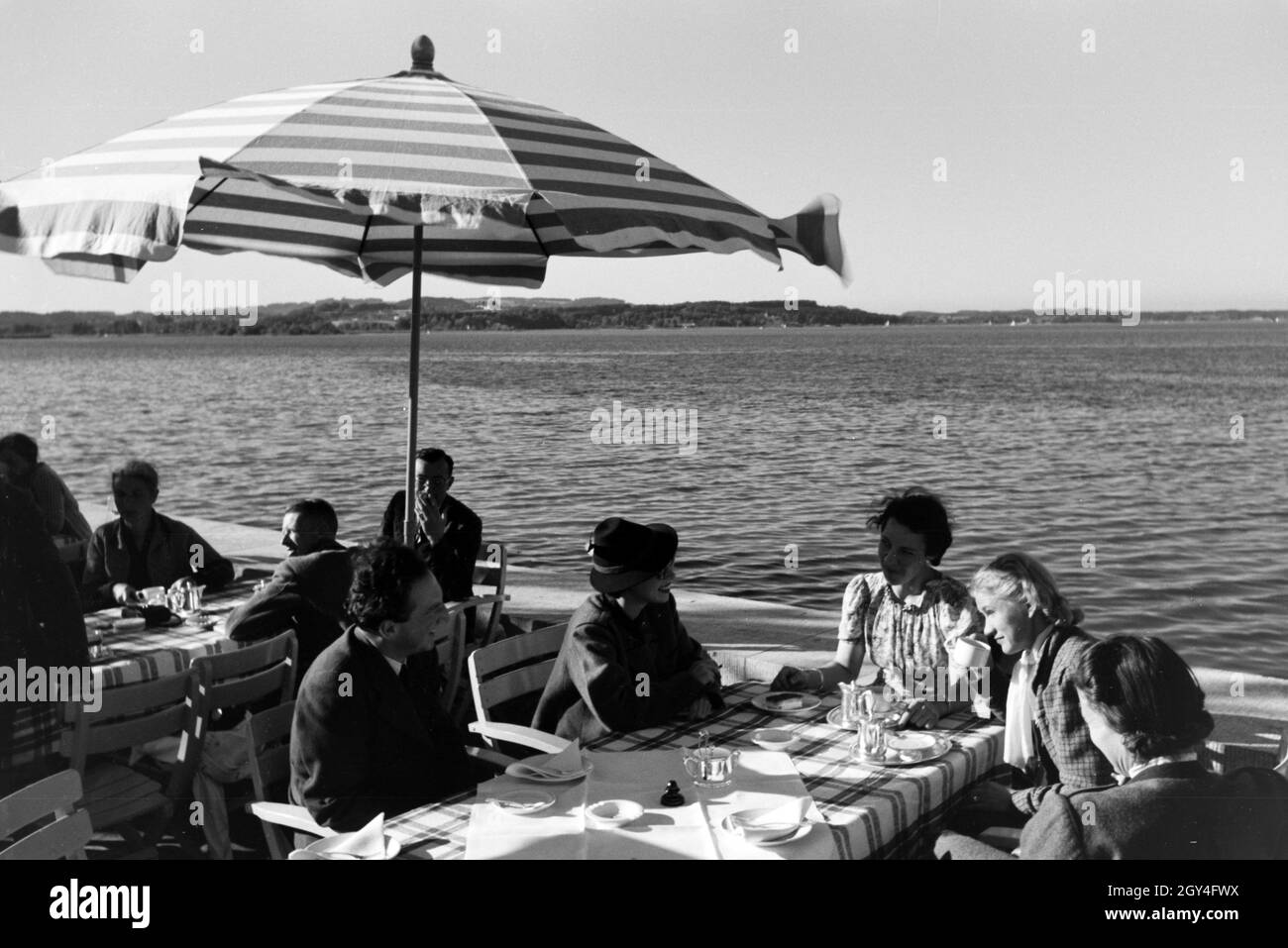 Gäste eines cafés am Chiemsee sitzen unter Sonnenschirmen auf der terrasse, Deutschland 1930er Jahre.Les clients d'un café du Chiemsee, assis sous des parasols sur la terrasse, Allemagne des années 1930. Banque D'Images