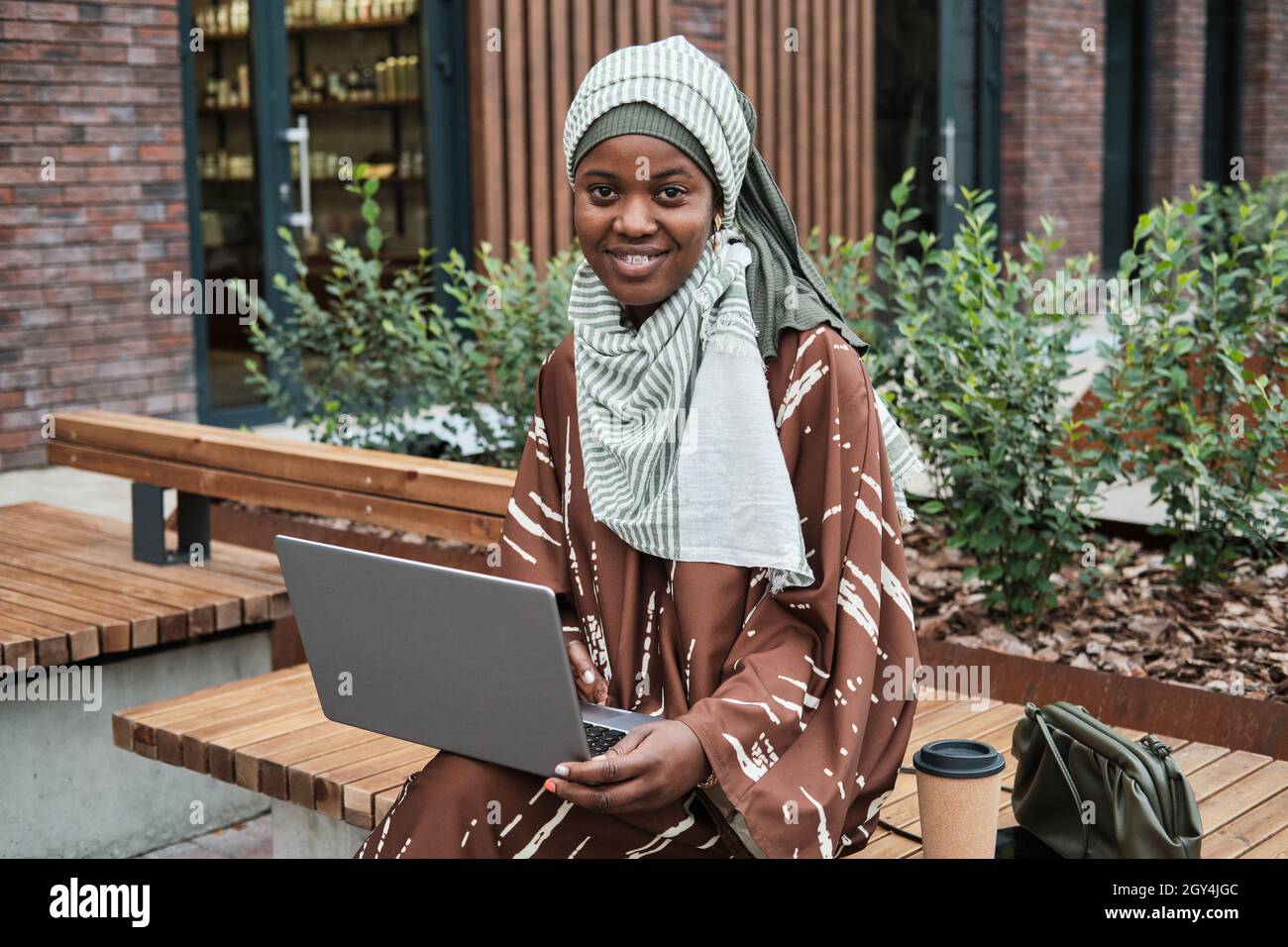 Portrait d'une femme d'affaires musulmane africaine assise sur le banc avec un ordinateur portable à l'extérieur et souriant à l'appareil photo Banque D'Images
