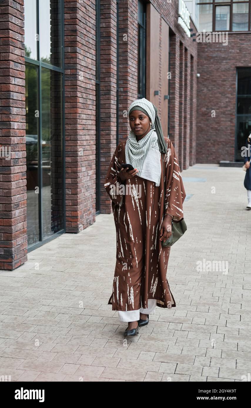 Jeune femme africaine vêtue de vêtements musulmans tenant un téléphone portable et marchant dans la rue à l'extérieur Banque D'Images