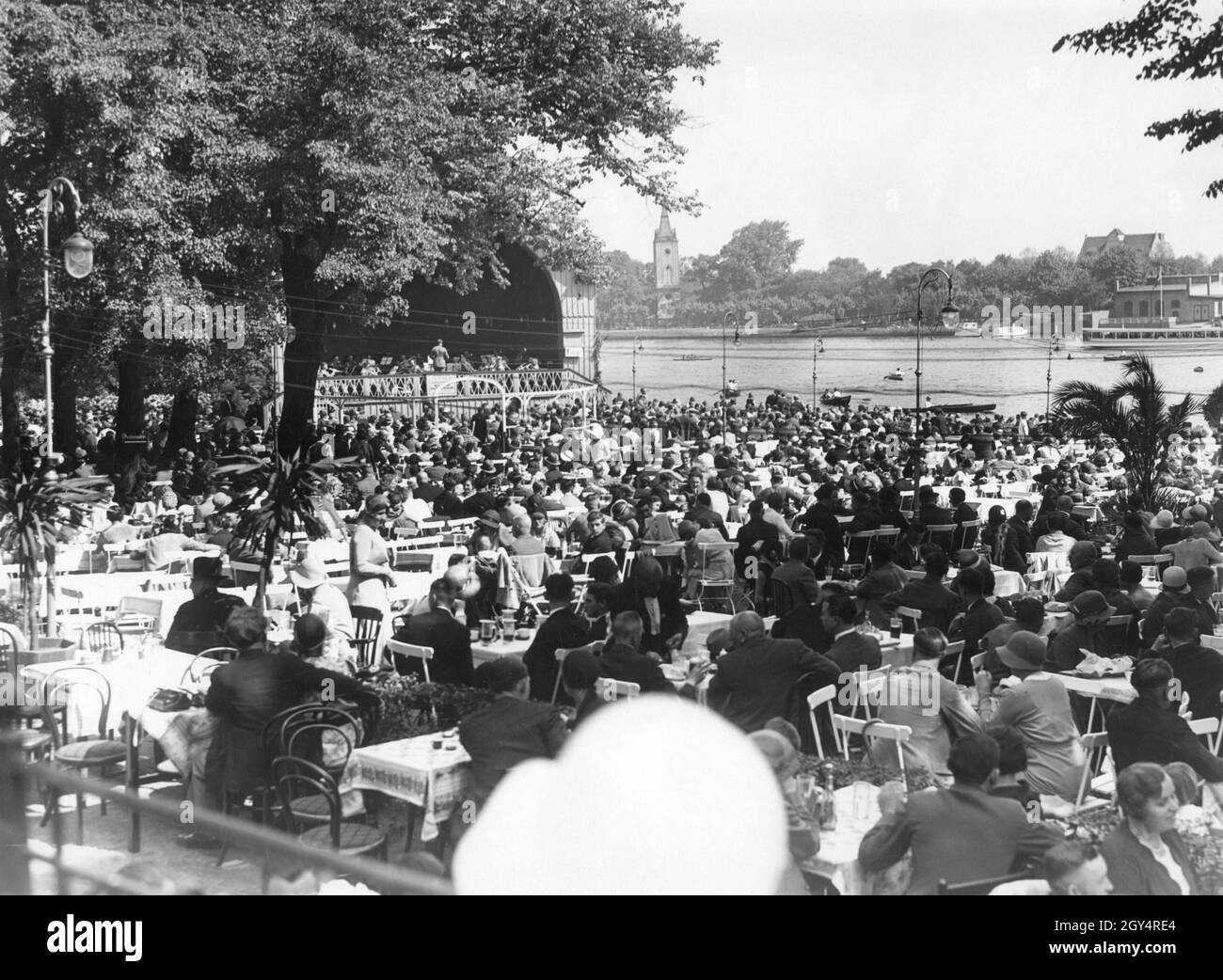 Lors d'une belle journée à l'été 1931, le restaurant Spreegarten du parc Treptower de Berlin est très fréquenté.Un orchestre donne un concert sur la scène.En arrière-plan, de l'autre côté de la Spree, on peut voir l'église du village de Stralau (au centre de l'image).[traduction automatique] Banque D'Images