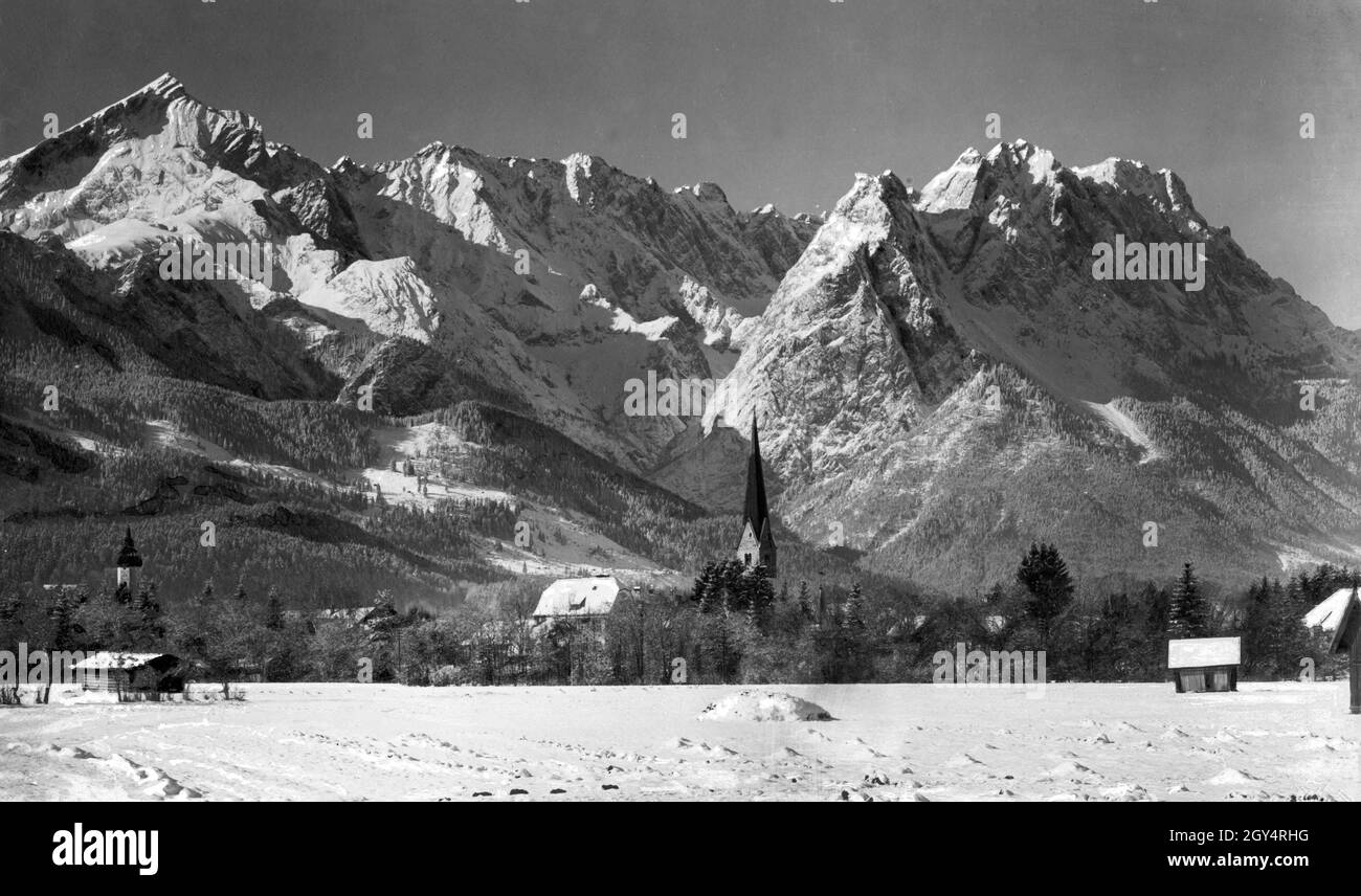 Les clochers des deux églises dans le quartier de Garmisch (St.Eglise paroissiale de Martin sur la gauche, Eglise paroissiale de Saint Martin sur la droite) se tenir dans la vallée enneigée d'hiver en face des sommets glacés des hautes montagnes (de gauche à droite: Alpspitze, Höllentalspitzen, Manndl, Waxenstein, Zugspitze).À gauche de la vieille église se trouve le nouveau bâtiment de l'école (Burgstraße 9, aujourd'hui une école élémentaire) construit dans le style baroque d'art nouveau.La photo du studio de Rudolf Rudolphi de Garmisch a été prise à l'hiver 1927.[traduction automatique] Banque D'Images