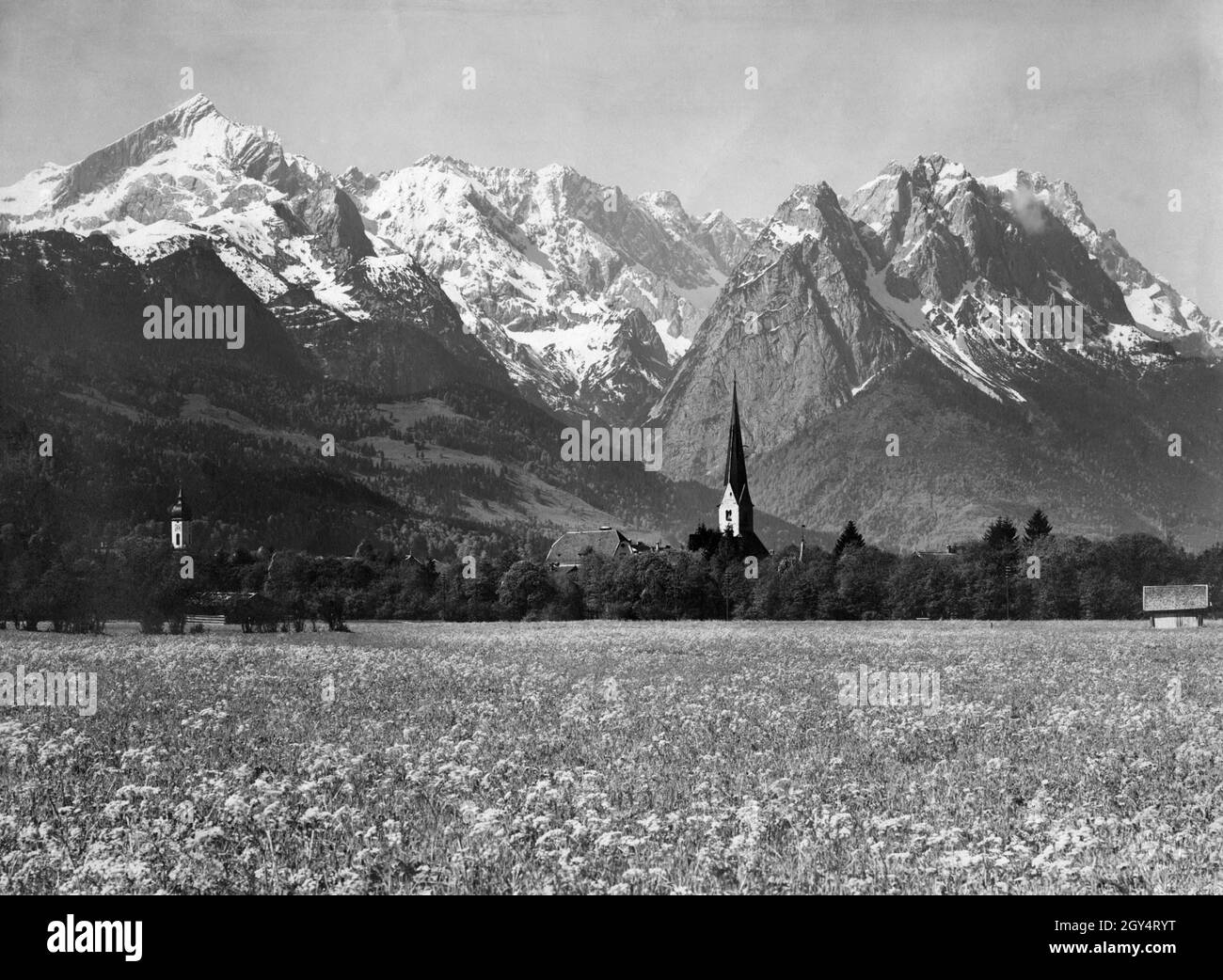 Les clochers des deux églises dans le quartier de Garmisch (St.Eglise paroissiale de Martin sur la gauche, l'ancienne église paroissiale de Saint Martin sur la droite) se trouve dans la vallée du début de l'été avec ses prairies fleuries devant les sommets encore enneigés des hautes montagnes (de gauche à droite : Alpspitze, Höllentalspitzen, Manndl, Waxenstein, Zugspitze).À gauche de la vieille église se trouve le nouveau bâtiment de l'école (Burgstraße 9, aujourd'hui une école élémentaire) construit dans le style baroque d'art nouveau.La photo du studio de Rudolf Rudolphi de Garmisch a été prise en 1928.[traduction automatique] Banque D'Images