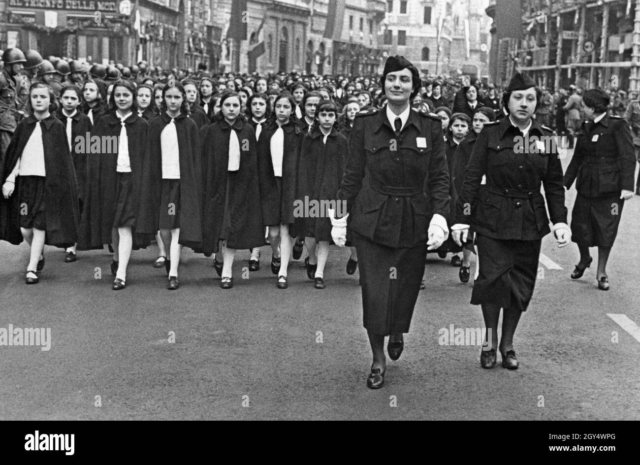 'Un groupe de filles, vêtues de hauts blancs et de cloques sombres, défilent dans une rue dans une ville italienne.Ils sont dirigés par deux chefs de groupe de la Gioventu Italiana del Littorio.Les filles appartiennent au groupe d'âge ''Piccole Italiane'', qui comprend les filles âgées de 8 à 14 ans.À gauche de la photo se trouvent les soldats de l'armée italienne.[traduction automatique]' Banque D'Images