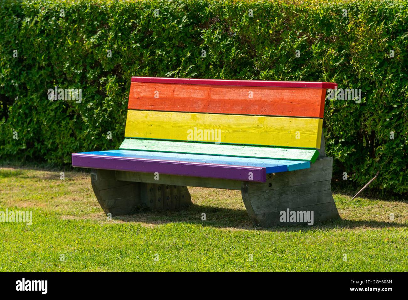 Un banc de parc peint gay Pride sur l'herbe verte et près d'une haie verte dans un parc.Le banc symbole les couleurs lgbt.Le coin salon en bois est de couleur. Banque D'Images