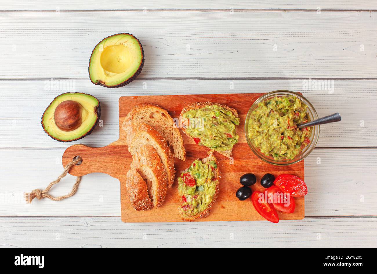 Plat Lay photo, guacamole fraîchement préparé dans un petit bol en verre, pain, tomates, olives à la table de travail et deux avocats à côté bureau en bois blanc. Banque D'Images