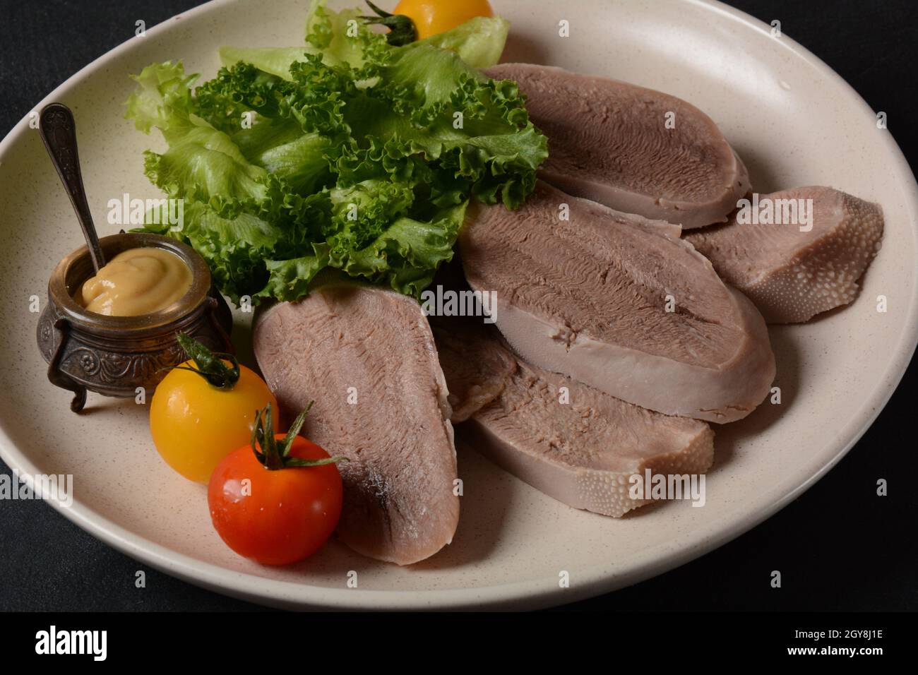 Tranches de langue de bœuf sur un plateau avec feuilles de laitue, tomates cerises et moutarde de Dijon sur fond noir Banque D'Images