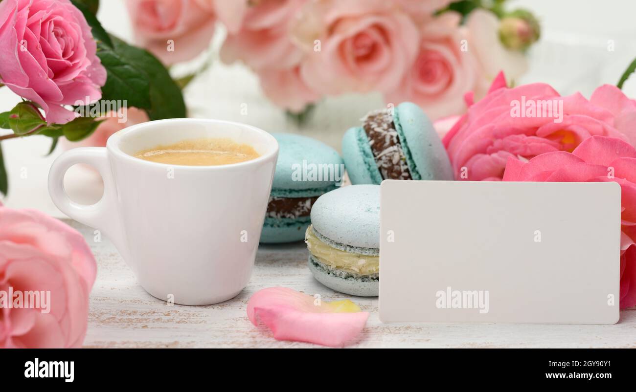 carte de visite blanche vierge avec café expresso et tasse en céramique blanche avec café et macaron bleu sur une table blanche, derrière un bouquet de r rose Banque D'Images