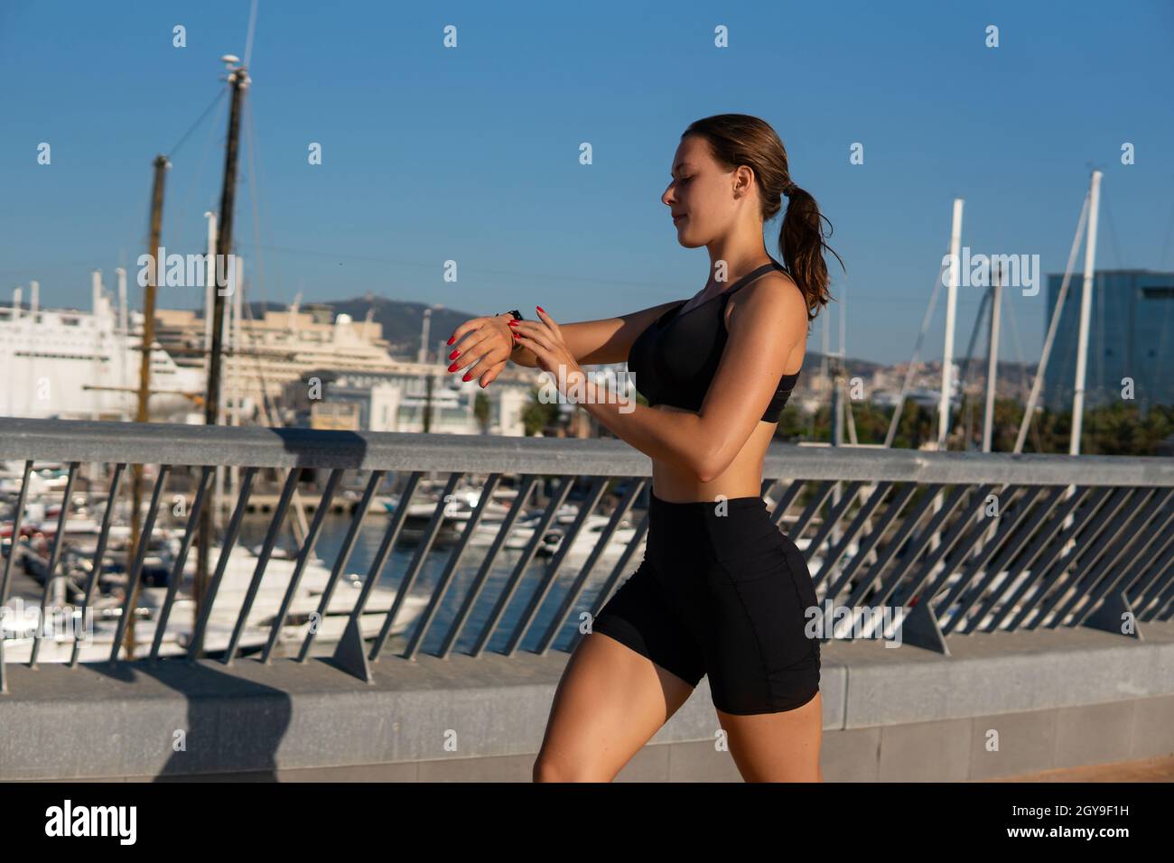 Faites équipe avec une athlète féminine dans un soutien-gorge et un short de sport en marchant sur un remblai et en vérifiant la montre intelligente pendant l'entraînement physique dans le port de la ville. Banque D'Images