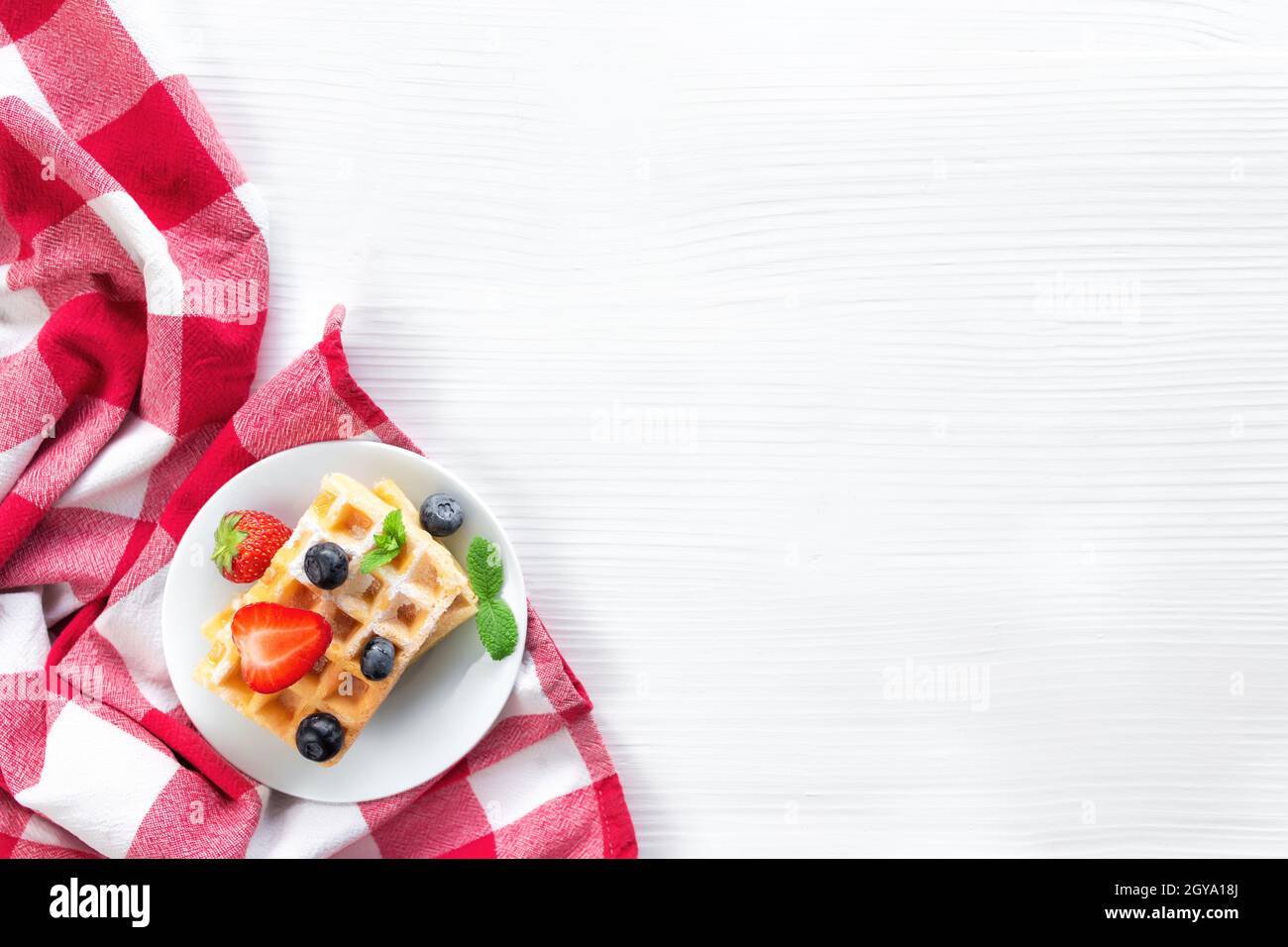 Assiette blanche sur serviette de cuisine rouge avec gaufres belges empilées faites maison, myrtille, fraises, feuille de menthe, sucre en poudre sur fond gris Banque D'Images