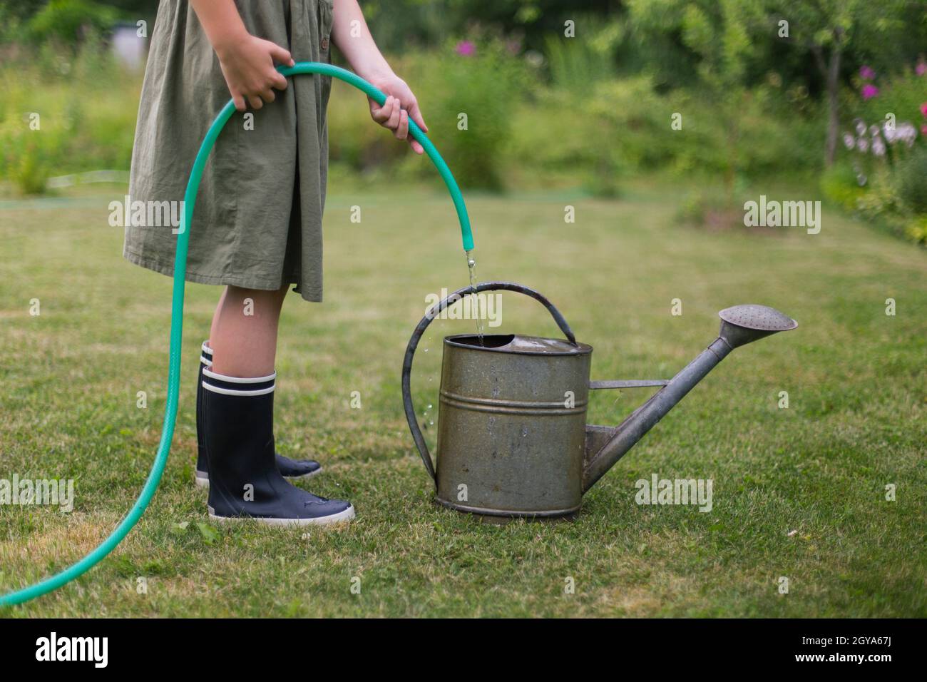 Un enfant en bottes de caoutchouc aspire l'eau d'un tuyau dans un arrosoir. Banque D'Images