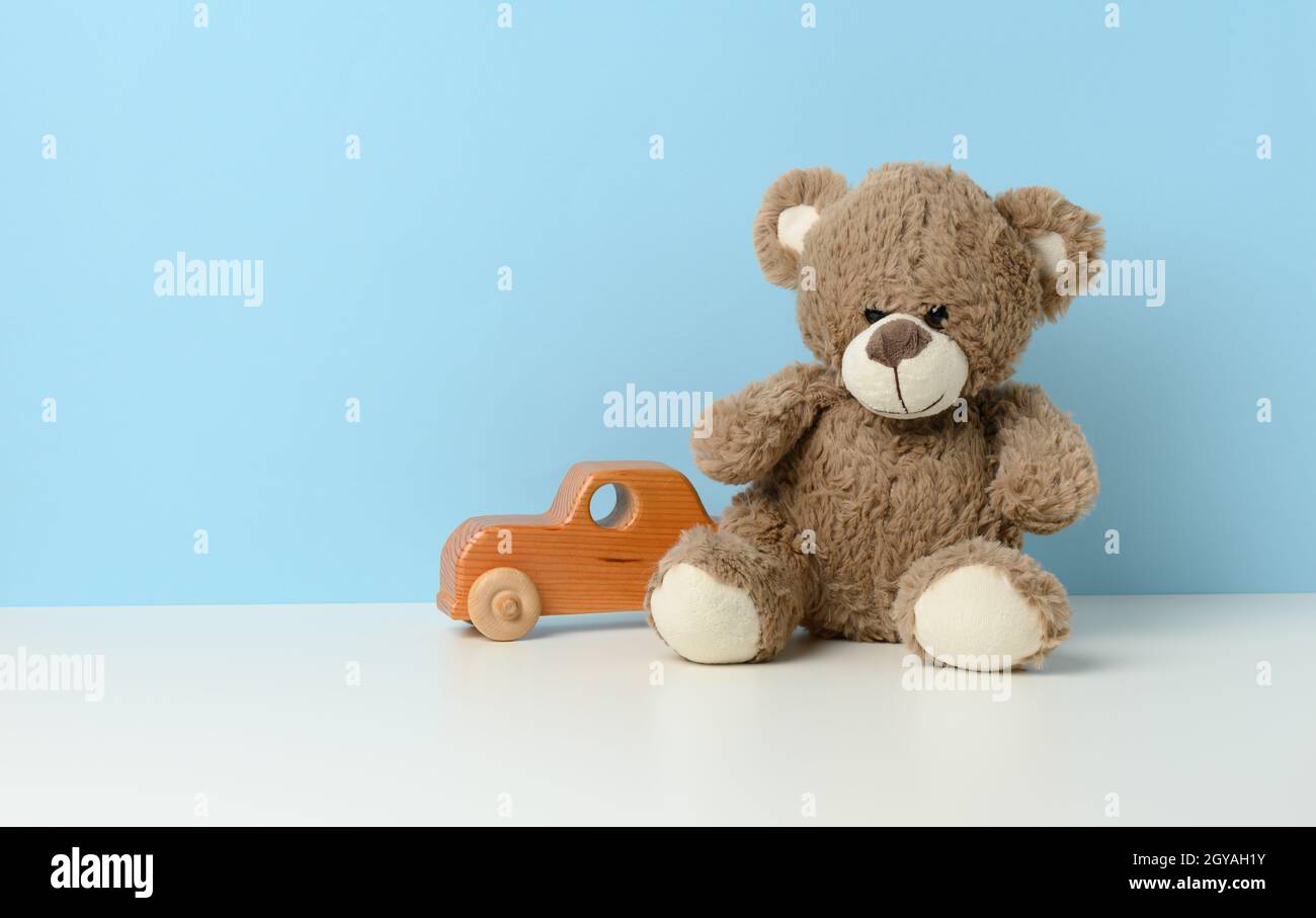 un adorable ours en peluche marron se trouve sur une table blanche et une voiture en bois pour enfants, sur fond bleu Banque D'Images