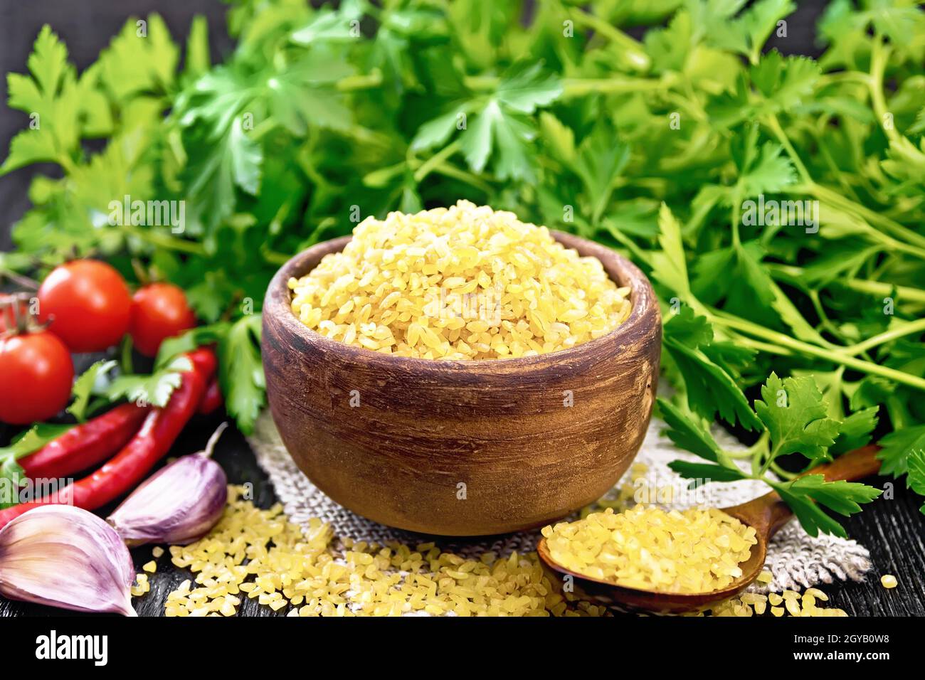 Gruaux bulgur - grains de blé cuits à la vapeur - dans un bol en argile et une cuillère sur le germe, les tomates, les poivrons chauds, l'ail et le persil sur fond de bois noir Banque D'Images