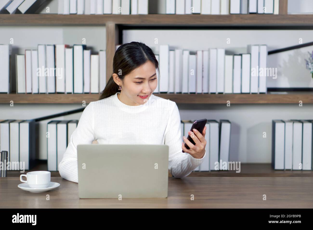 Jeune femme asiatique lisant un message texte sur un téléphone portable tout en faisant sa thèse sur un ordinateur portable dans la bibliothèque. Une tasse de café a été placée sur l'onglet Banque D'Images