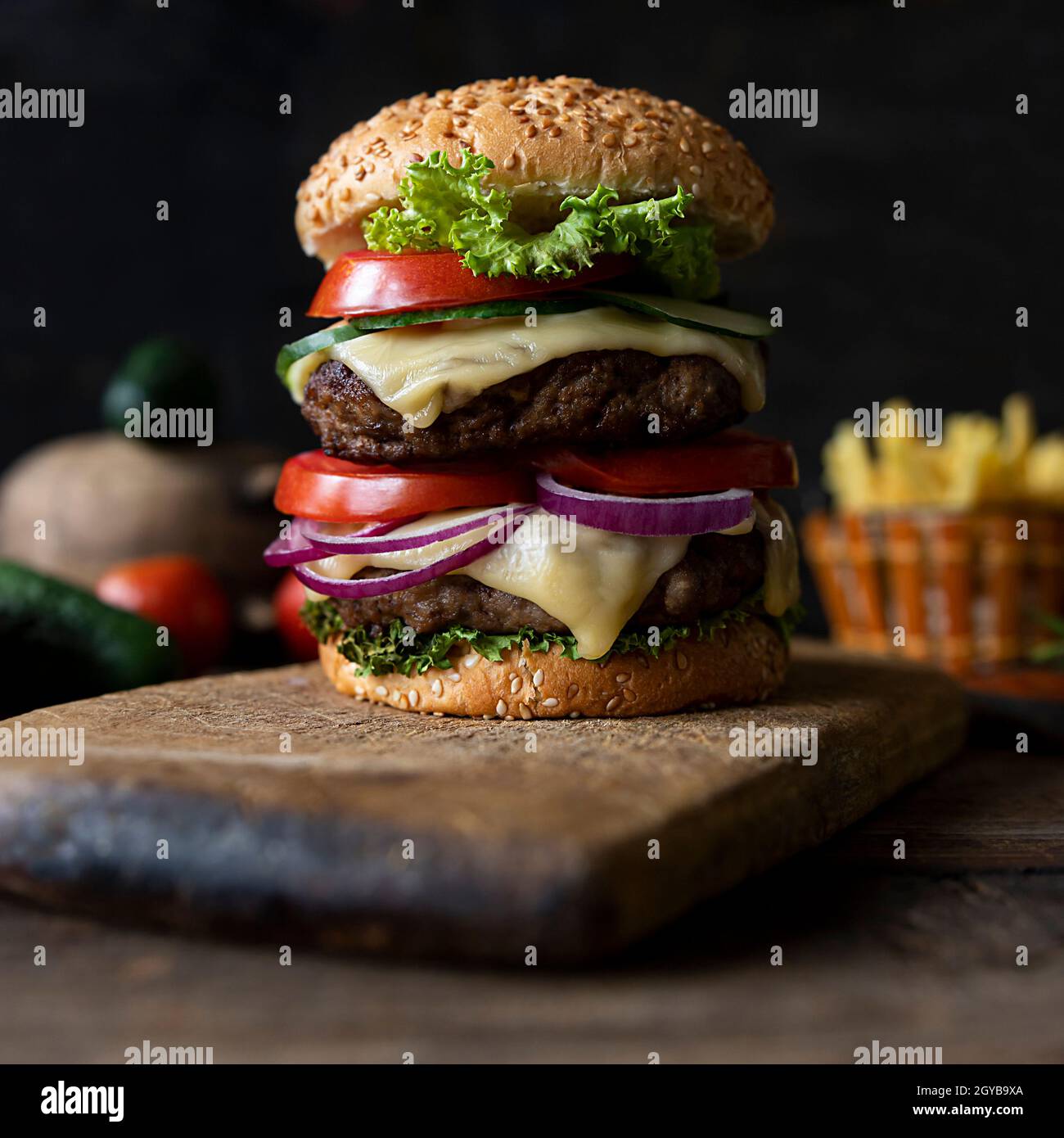 Hamburger aux tomates, oignons, concombre, laitue et fromage fondu servi sur un panneau de bois rustique avec frites et légumes en arrière-plan Banque D'Images