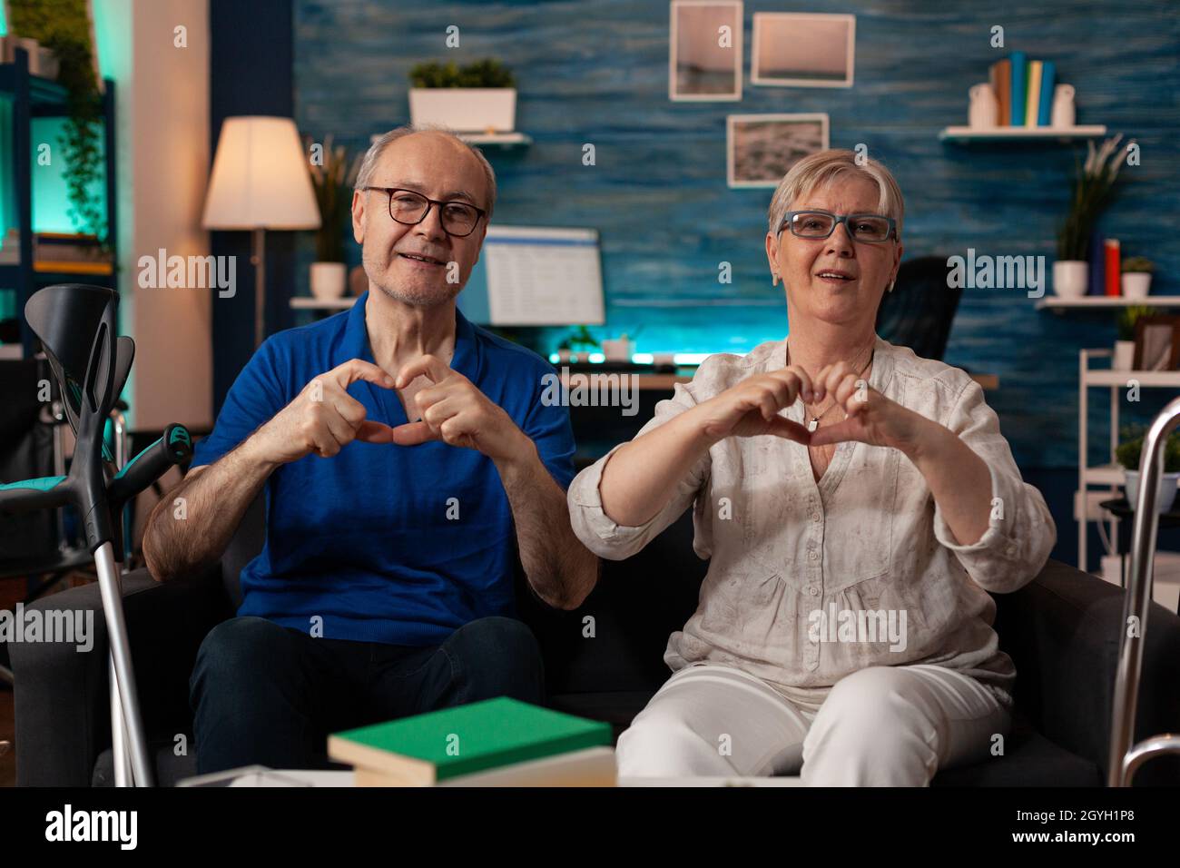 Couple âgé créant la forme du cœur avec les mains tout en regardant la caméra dans le salon.Les gens mariés amoureux font le symbole de l'affection ensemble et assis sur un canapé à la maison.Relation senior Banque D'Images