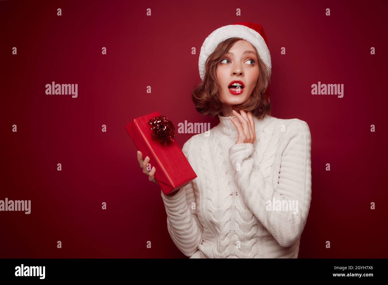 Femme rêveuse en chapeau de père Noël avec cadeau sur fond rouge Banque D'Images