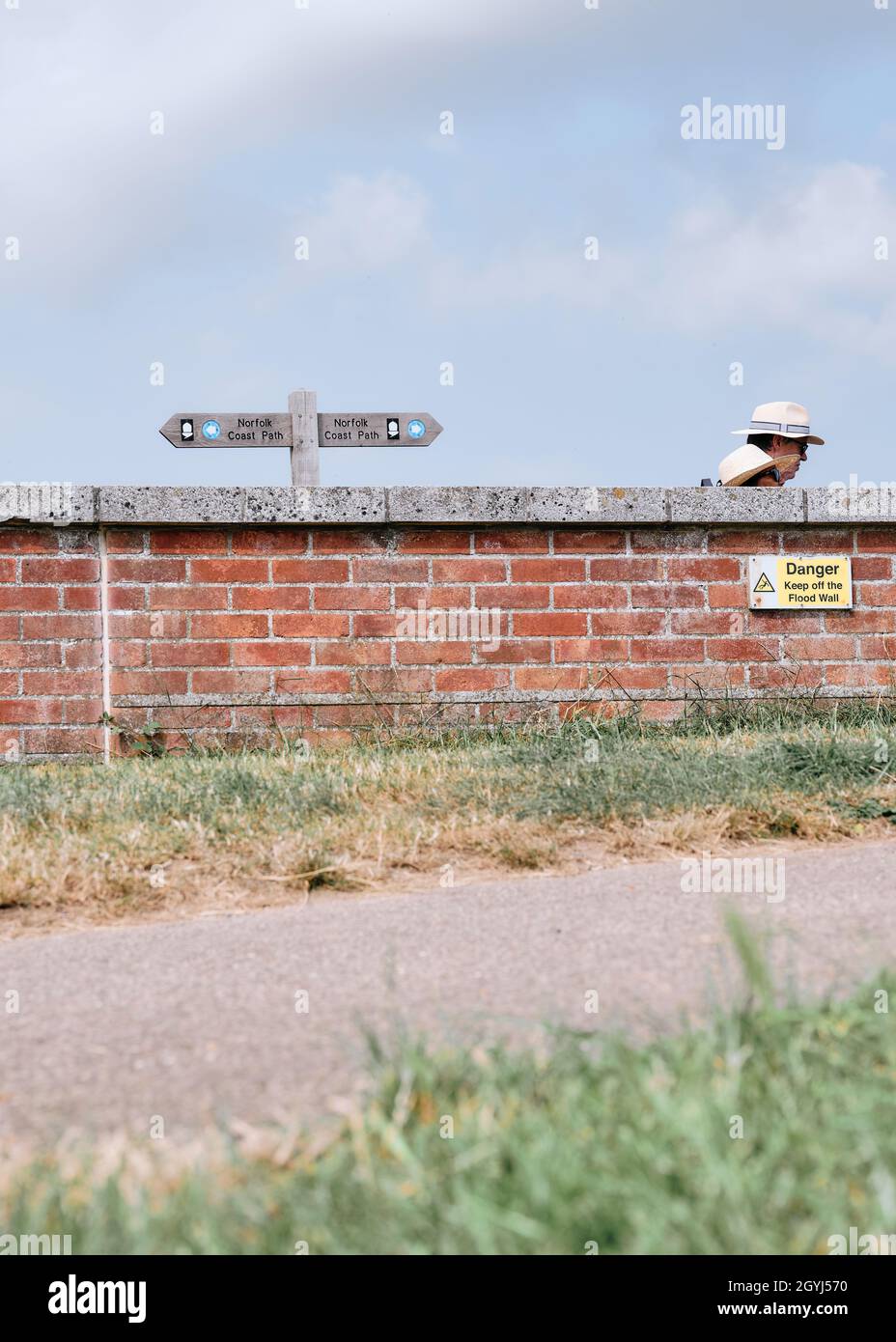 Une vue étrange sur le Norfolk Coast Path avec un panneau et des marcheurs portant des chapeaux de soleil à moitié cachés par un mur de défense contre les inondations. Banque D'Images