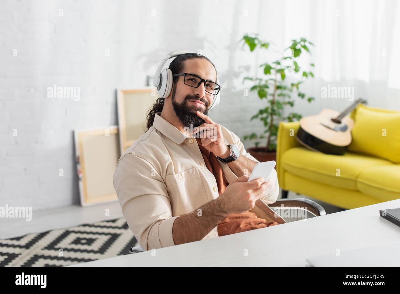homme hispanique rêveur et souriant avec un smartphone qui écoute de la musique dans un casque à la maison Banque D'Images