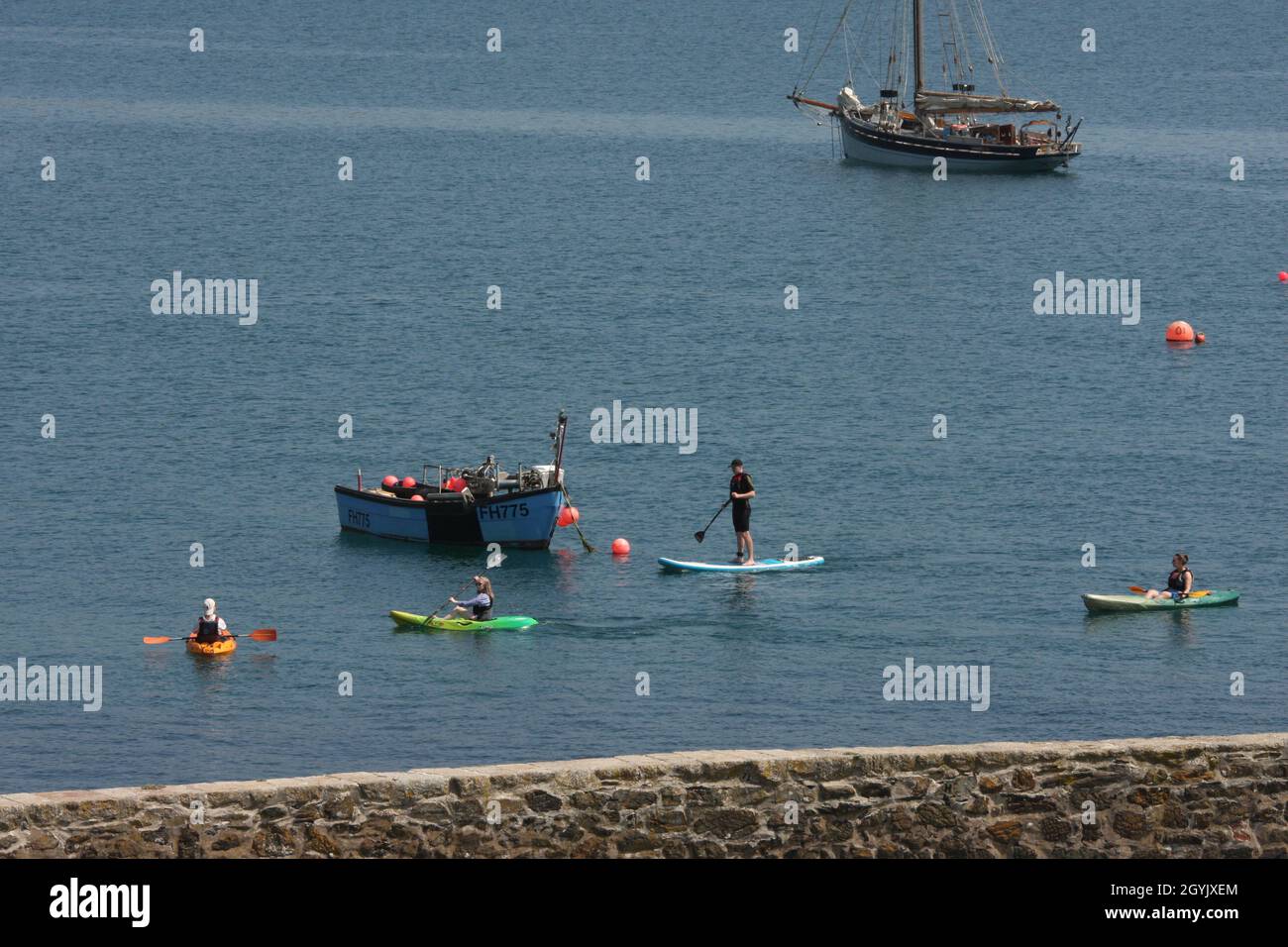 Les gens pagayent dans des canots et paddleboard devant un petit bateau de pêche ancré et un vieux yacht en bois, Cornwall, Royaume-Uni Banque D'Images