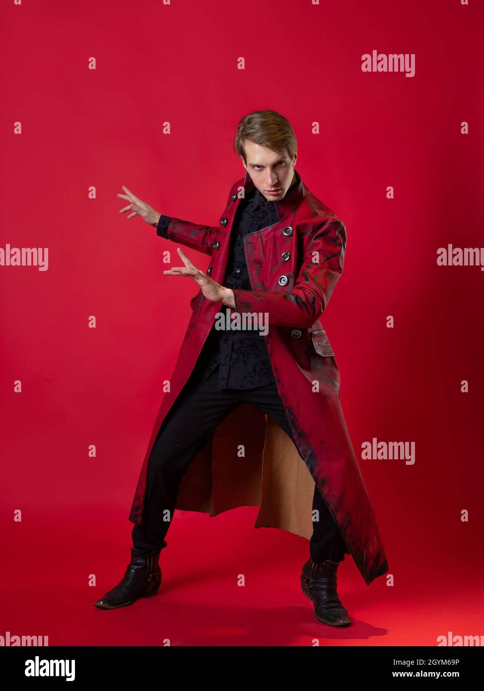 Un jeune homme scandaleux dans un manteau rouge audacieux dans un style noir vintage, pose épique et fait un geste de main. Photo sur fond rouge Banque D'Images