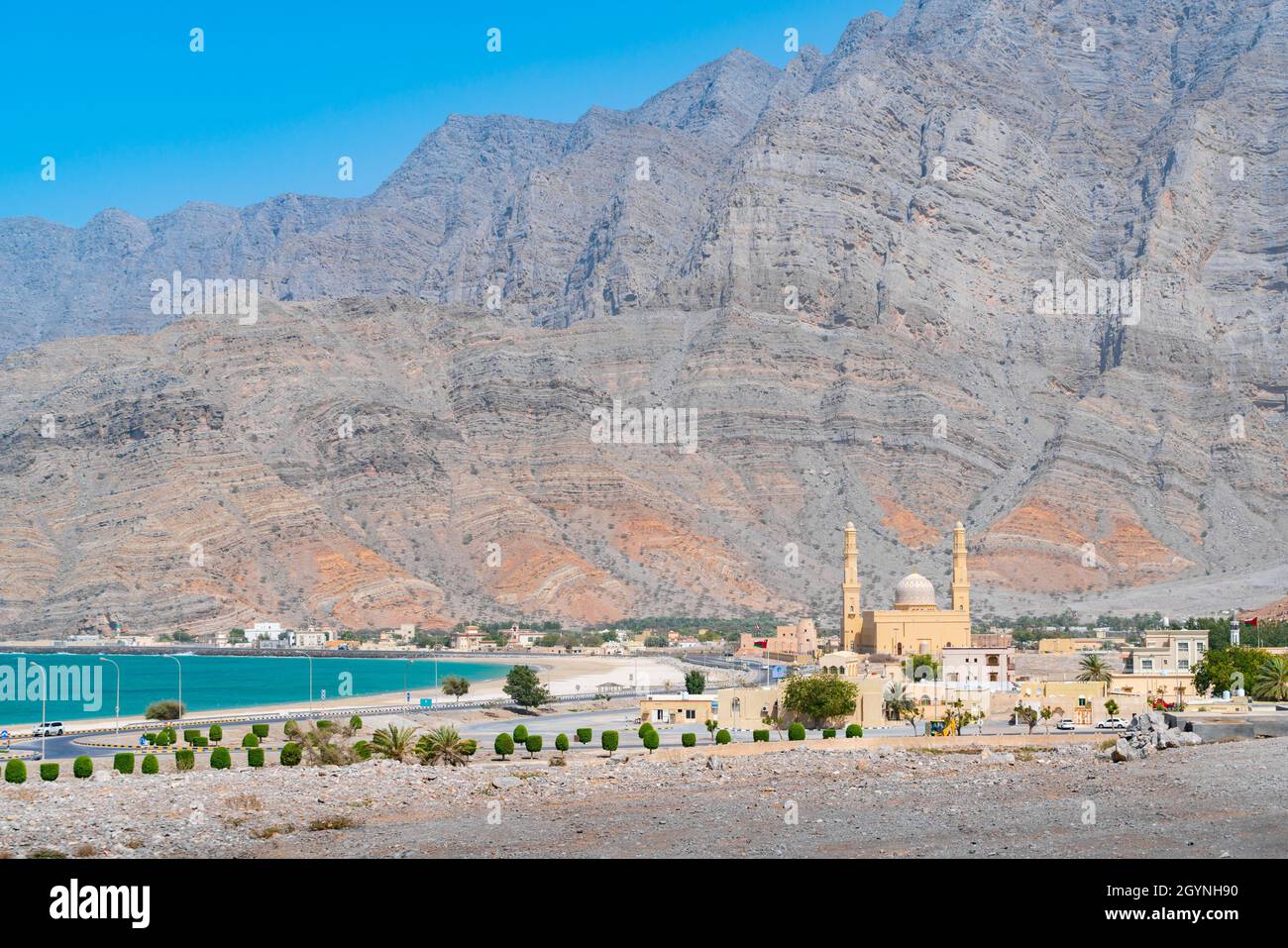 Mosquée avec deux minarets et un petit fort avec l'océan et de hautes falaises de montagne en arrière-plan lors d'une chaude journée ensoleillée.Voyage dans la péninsule de Musandam, Oman Banque D'Images