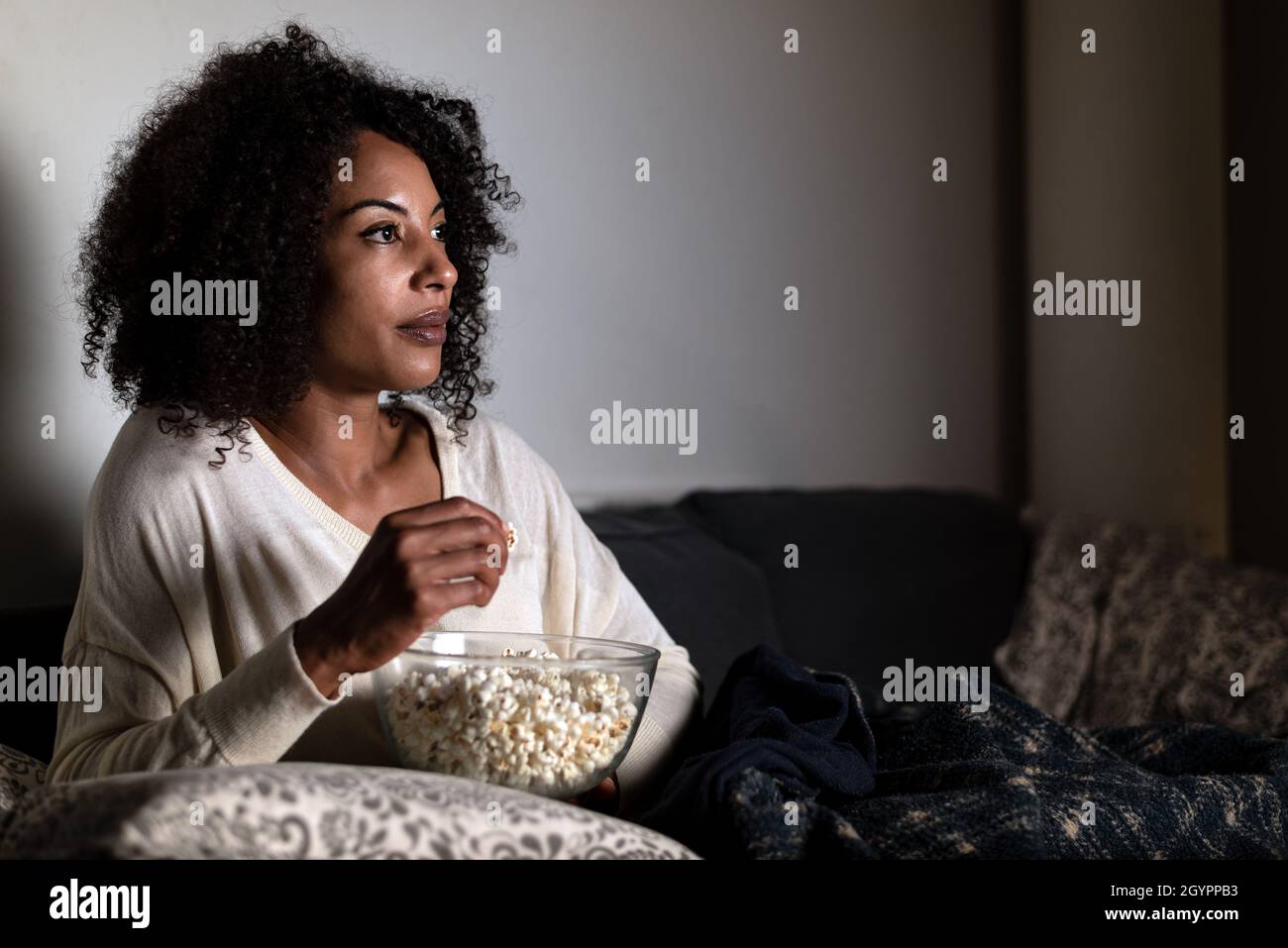 portrait horizontal d'une femme afro-américaine assise sur un canapé.Le visage est éclairé par la lumière du téléviseur.Elle tient un bol de pop-corn Banque D'Images