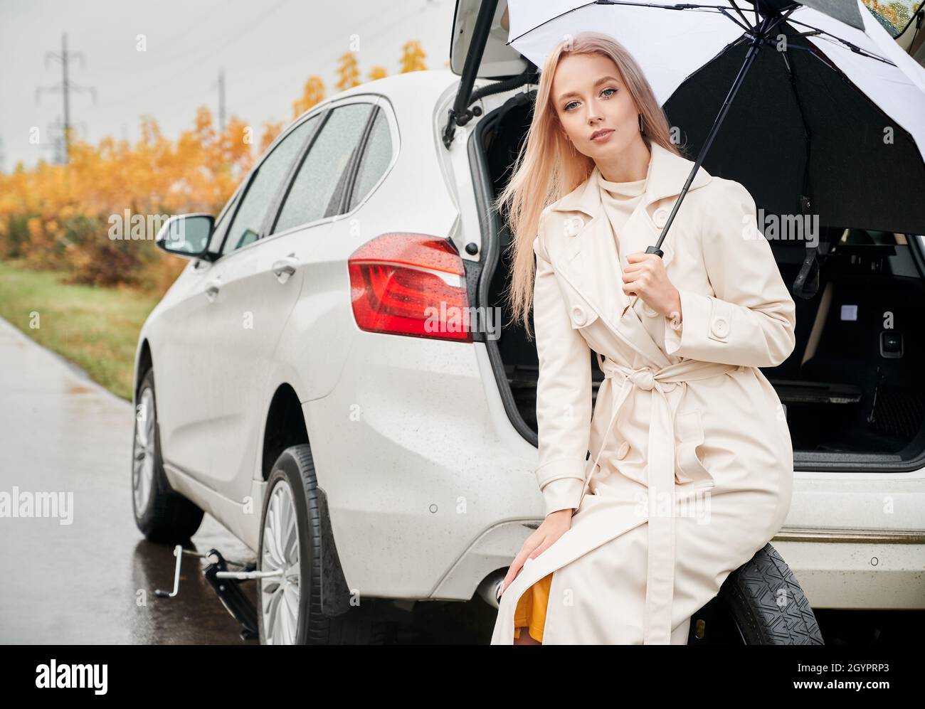Portrait d'une jolie femme d'affaires blonde assise sur la roue de secours, tenant le parapluie de la pluie.Sur l'arrière-plan, voiture blanche avec coffre ouvert et buissons jaunes d'automne. Banque D'Images