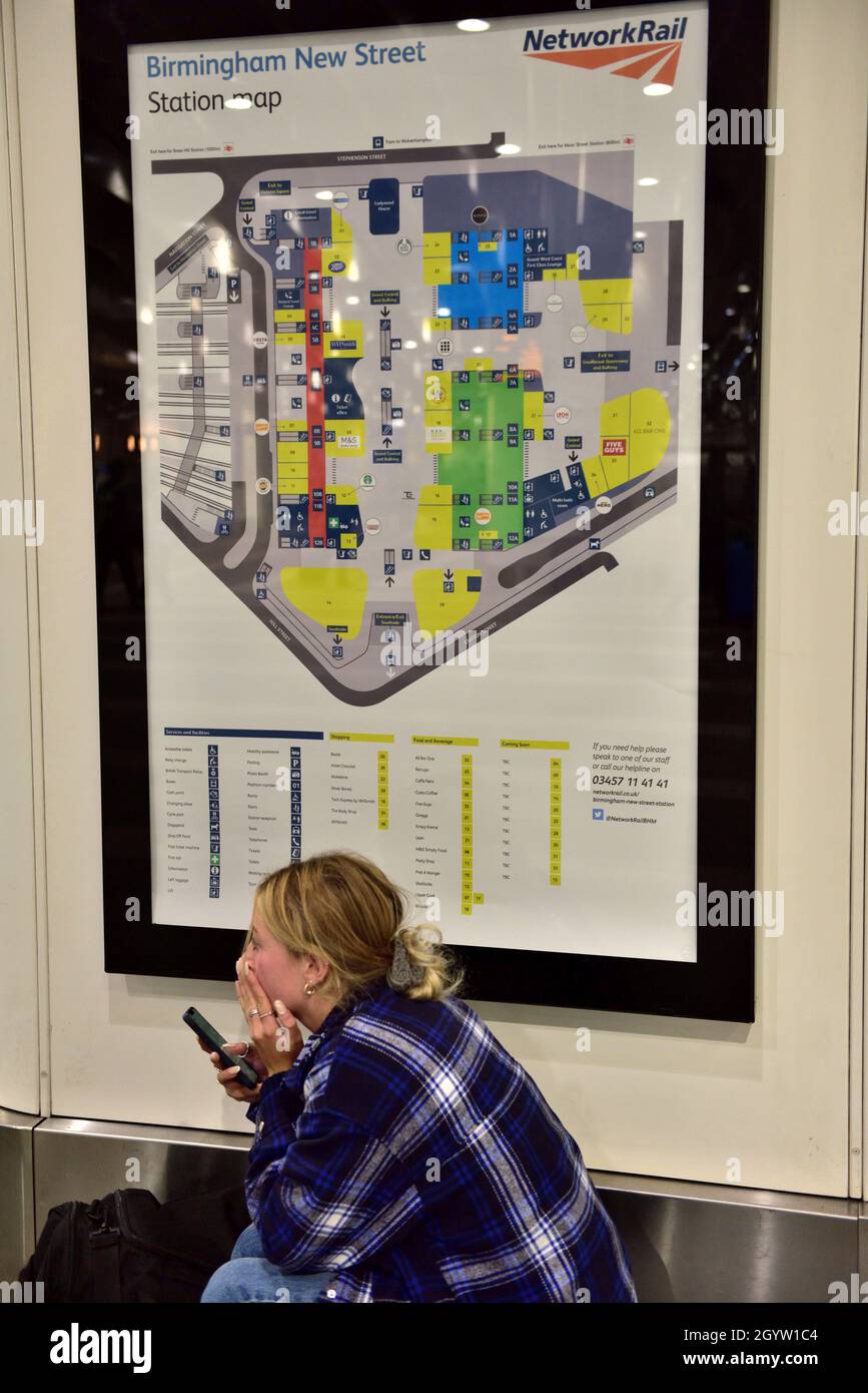 Jeune femme assise avec un téléphone au-dessous de la carte de la gare de Birmingham New Street Banque D'Images