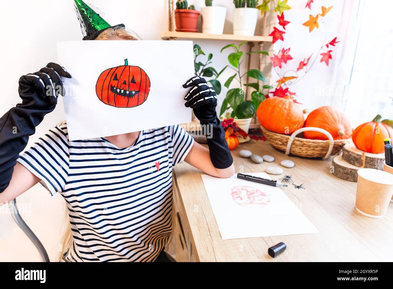 Un enfant montre son dessin pour Halloween, assis à un bureau près de la fenêtre, entouré de citrouilles et de plantes de maison. Dessin d'un rire orange Banque D'Images
