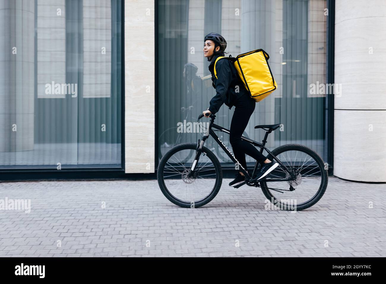 Vue latérale d'une jeune femme à vélo dans la ville.Coursier avec un sac à dos thermique en son chemin pour livrer la nourriture à un client. Banque D'Images