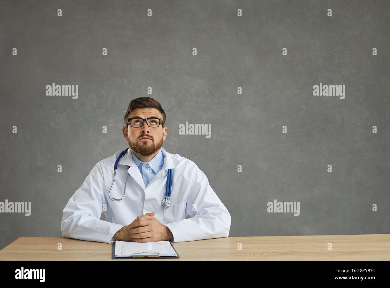 Un jeune médecin masculin rêveur avec une expression pensive regarde assis à une table sur un fond gris. Banque D'Images