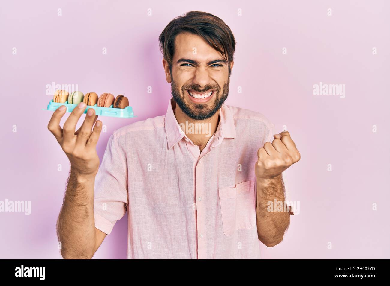 Un jeune hispanique qui tient de délicieuses pâtisseries de macrons criant fièrement, célébrant la victoire et le succès très excité avec le bras relevé Banque D'Images